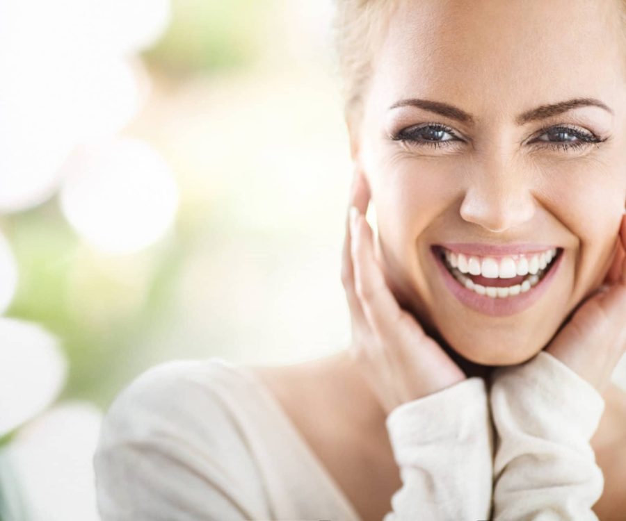 Closeup portrait of an attractive mid 30's woman gently touching her neck and smiling while looking at the camera and sincerely smiling. Blurry green background.