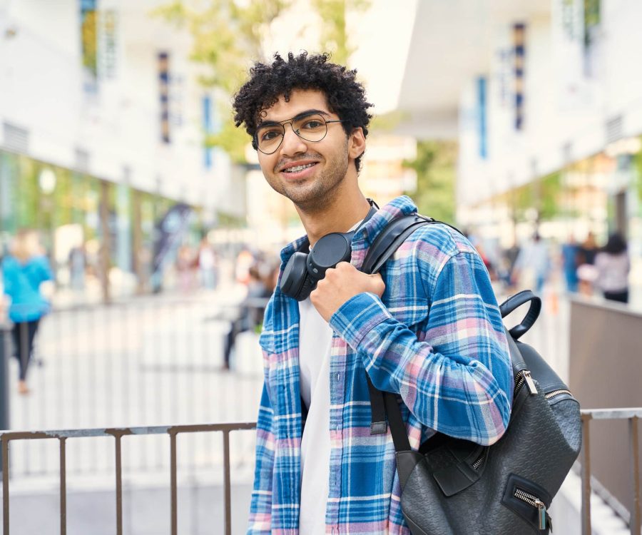 Young curly middle eastern man smiling happy while standing with backpack and using headphones at the city. Urban lifestyle concept