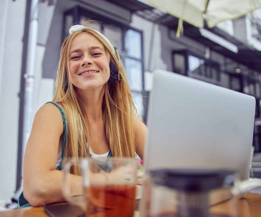 Joyful woman with freckles is sitting at the table in a cafe with a laptop and a cup of tea looking at the camera